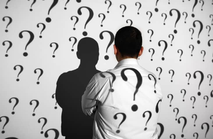 Businessman standing in front of projection screen. Projector reflects questions marks to screen and businessman's back.
